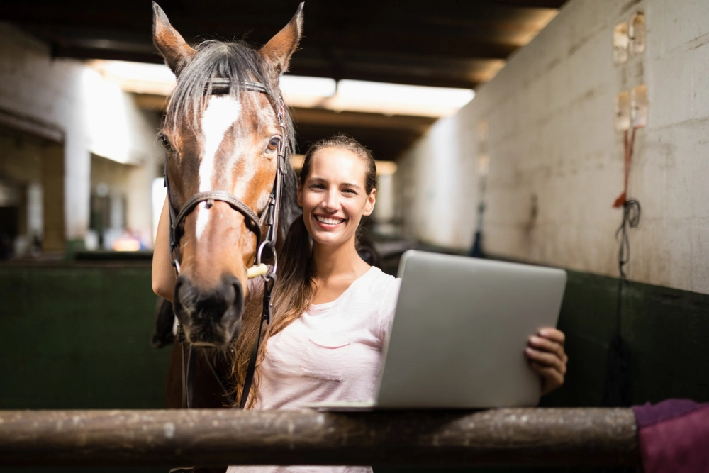 Woman holding laptop beside horse in stable